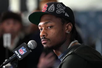 Stefon Diggs - Ein Mann mit schwarzer Mütze und Kapuzenpulli, der Stefon Diggs ähnelt, sitzt während einer Presseveranstaltung an einem NFL-Mikrofon und schaut über seine Schulter in die Kamera. Alt-Text wurde KI-generiert.