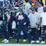 Football-Trainer Klint Kubiak steht mit einem Spielbuch an der Seitenlinie, umgeben von Spielern und Mitarbeitern in Seattle Seahawks-Kleidung während eines Spiels gegen die Las Vegas Raiders. Alt-Text wurde KI-generiert.