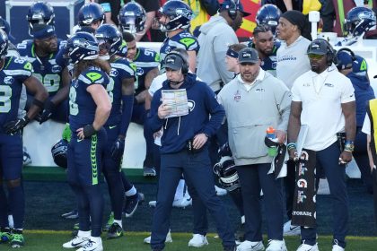 Football-Trainer Klint Kubiak steht mit einem Spielbuch an der Seitenlinie, umgeben von Spielern und Mitarbeitern in Seattle Seahawks-Kleidung während eines Spiels gegen die Las Vegas Raiders. Alt-Text wurde KI-generiert.