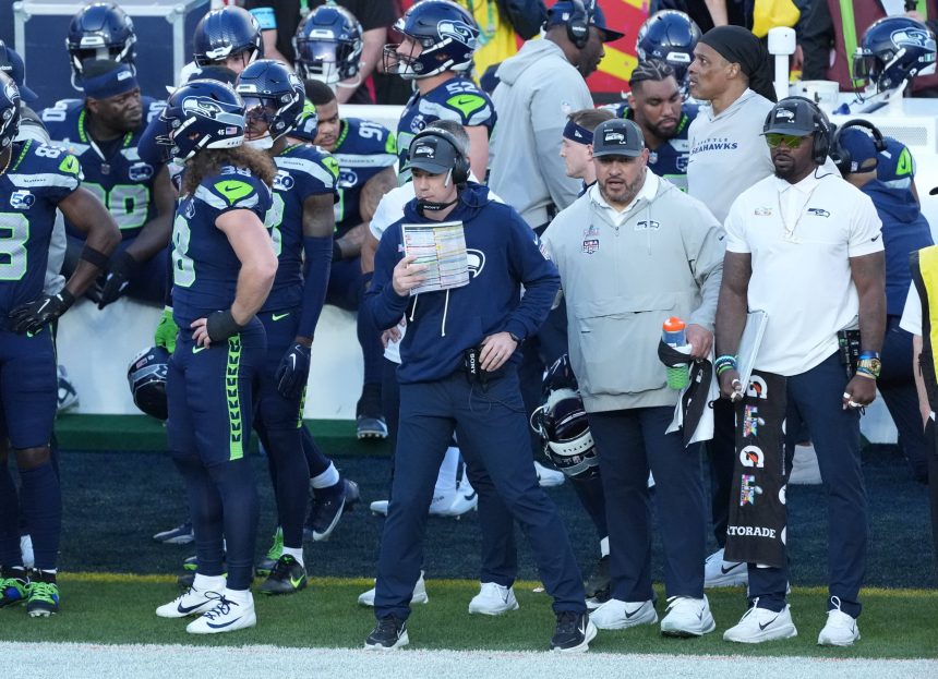 Football-Trainer Klint Kubiak steht mit einem Spielbuch an der Seitenlinie, umgeben von Spielern und Mitarbeitern in Seattle Seahawks-Kleidung während eines Spiels gegen die Las Vegas Raiders. Alt-Text wurde KI-generiert.