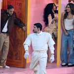 Bad Bunny - Super Bowl LX - Eine Gruppe von Menschen in hellen Outfits (darunter Jessica Alba, Cardi B, Karol G)steht und interagiert auf einem rosafarbenen Bühnenbild mit Türen und Säulen, das an eine lebendige Halftime-Show-Performance mit versteckten Botschaften erinnert, die das Publikum entdecken kann. Alt-Text wurde KI-generiert.