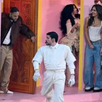 Bad Bunny - Super Bowl LX - Eine Gruppe von Menschen in hellen Outfits (darunter Jessica Alba, Cardi B, Karol G)steht und interagiert auf einem rosafarbenen Bühnenbild mit Türen und Säulen, das an eine lebendige Halftime-Show-Performance mit versteckten Botschaften erinnert, die das Publikum entdecken kann. Alt-Text wurde KI-generiert.