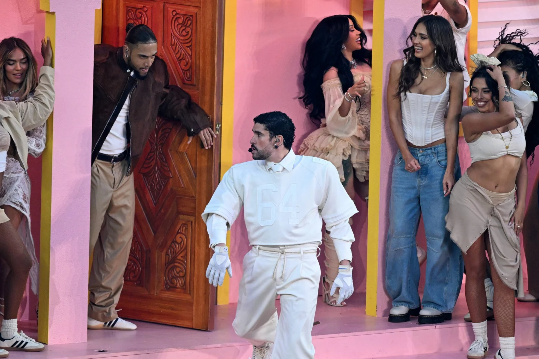 Bad Bunny - Super Bowl LX - Eine Gruppe von Menschen in hellen Outfits (darunter Jessica Alba, Cardi B, Karol G)steht und interagiert auf einem rosafarbenen Bühnenbild mit Türen und Säulen, das an eine lebendige Halftime-Show-Performance mit versteckten Botschaften erinnert, die das Publikum entdecken kann. Alt-Text wurde KI-generiert.