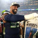 Ein Spieler der Seattle Seahawks in Uniform lächelt und hält eine "CHAMPS"-Zeitung auf dem Spielfeld nach dem Super Bowl LX in der Hand. Im Hintergrund sind die Lichter des Stadions und jubelnde Fans zu sehen, die ihren großen Sieg feiern. Alt-Text wurde KI-generiert.