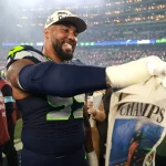 Ein Spieler der Seattle Seahawks in Uniform lächelt und hält eine "CHAMPS"-Zeitung auf dem Spielfeld nach dem Super Bowl LX in der Hand. Im Hintergrund sind die Lichter des Stadions und jubelnde Fans zu sehen, die ihren großen Sieg feiern. Alt-Text wurde KI-generiert.