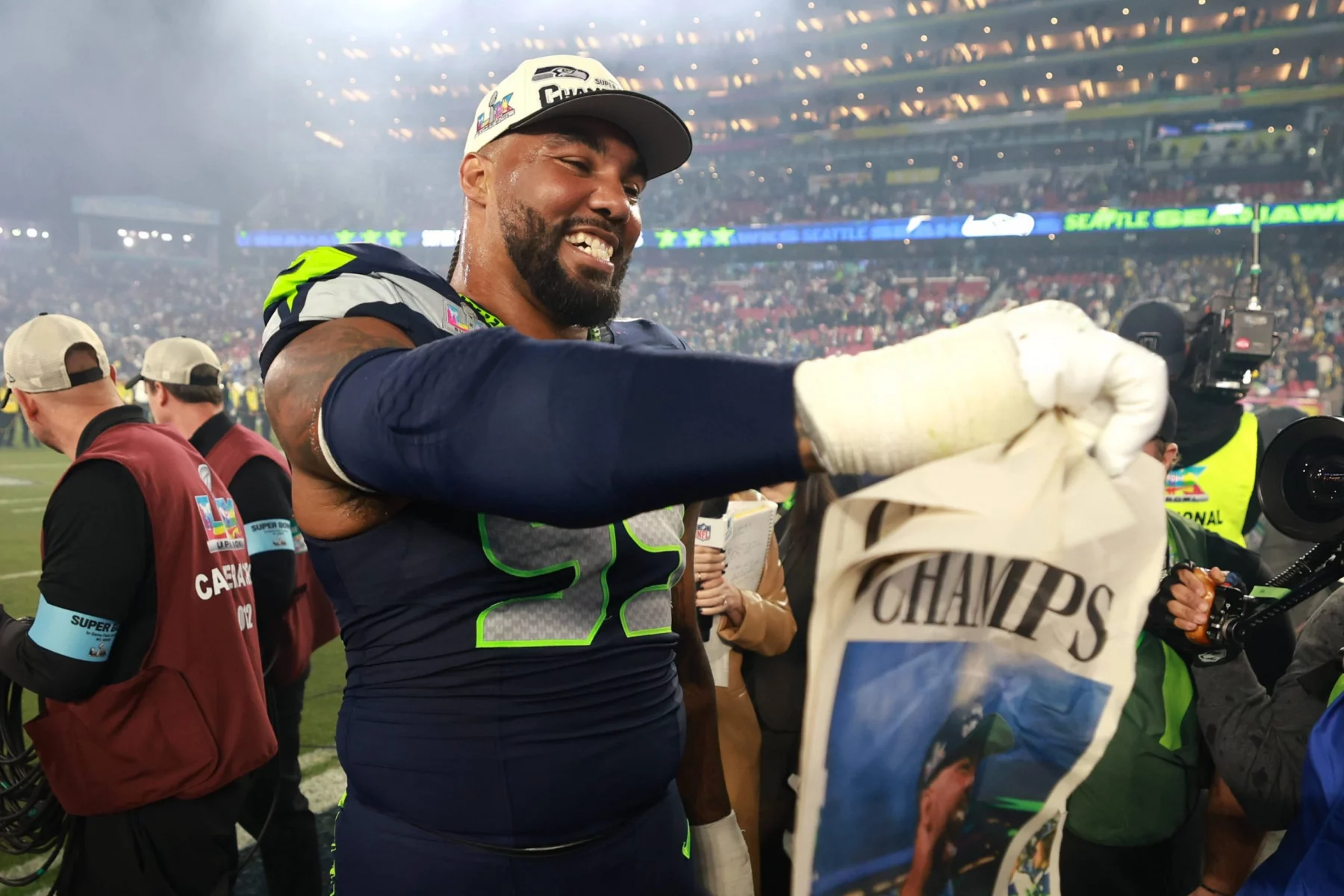 Ein Spieler der Seattle Seahawks in Uniform lächelt und hält eine "CHAMPS"-Zeitung auf dem Spielfeld nach dem Super Bowl LX in der Hand. Im Hintergrund sind die Lichter des Stadions und jubelnde Fans zu sehen, die ihren großen Sieg feiern. Alt-Text wurde KI-generiert.