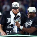 Sam Darnold und Kenneth Walker III - Zwei Football-Spieler mit "Champions"-Hüten und -Shirts lächeln, als sie während der Super Bowl 50-Feier die Vince Lombardi Trophy halten und damit beweisen, dass alles möglich ist, wenn du an dich selbst glaubst. Alt-Text wurde KI-generiert.