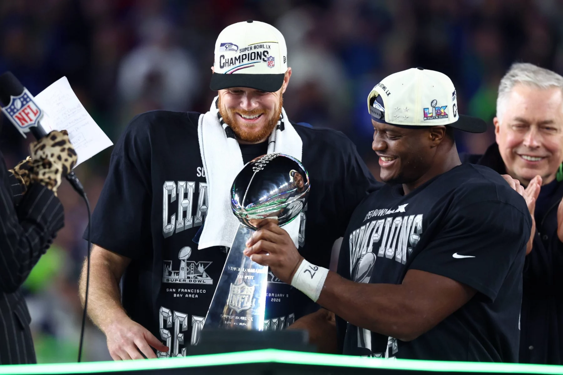 Sam Darnold und Kenneth Walker III - Zwei Football-Spieler mit "Champions"-Hüten und -Shirts lächeln, als sie während der Super Bowl 50-Feier die Vince Lombardi Trophy halten und damit beweisen, dass alles möglich ist, wenn du an dich selbst glaubst. Alt-Text wurde KI-generiert.