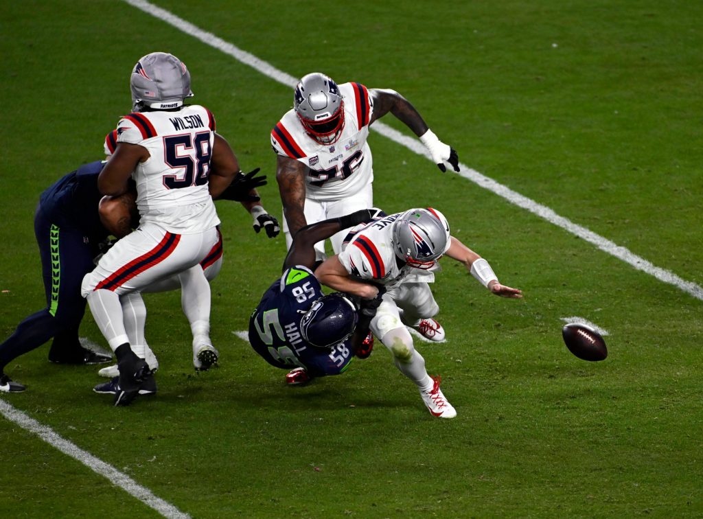 Drake Maye - Ein Footballspieler in Weiß wird von einem Spieler in Marineblau umgerannt, wodurch der Ball losfliegt, während die anderen Spieler zuschauen. Das erinnert an die Intensität, die man nach einer Super-Bowl-Niederlage bei einem Spiel auf einem grünen Feld sieht. Alt-Text wurde KI-generiert.