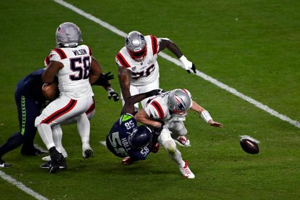 Drake Maye - Ein Footballspieler in Weiß wird von einem Spieler in Marineblau umgerannt, wodurch der Ball losfliegt, während die anderen Spieler zuschauen. Das erinnert an die Intensität, die man nach einer Super-Bowl-Niederlage bei einem Spiel auf einem grünen Feld sieht. Alt-Text wurde KI-generiert.