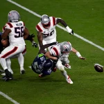 Drake Maye - Ein Footballspieler in Weiß wird von einem Spieler in Marineblau umgerannt, wodurch der Ball losfliegt, während die anderen Spieler zuschauen. Das erinnert an die Intensität, die man nach einer Super-Bowl-Niederlage bei einem Spiel auf einem grünen Feld sieht. Alt-Text wurde KI-generiert.