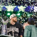 Sam Darnold - Seattle Seahawks - Ein Football-Spieler, der ein "Champion"-Shirt und eine Kappe trägt, steht neben der Super-Bowl-Trophäe, während Konfetti herabfällt. Um ihn herum auf dem Spielfeld feiern andere den Sieg der Seattle Seahawks. Alt-Text wurde KI-generiert.