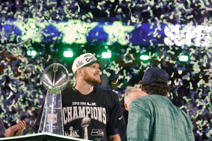 Sam Darnold - Seattle Seahawks - Ein Football-Spieler, der ein "Champion"-Shirt und eine Kappe trägt, steht neben der Super-Bowl-Trophäe, während Konfetti herabfällt. Um ihn herum auf dem Spielfeld feiern andere den Sieg der Seattle Seahawks. Alt-Text wurde KI-generiert.