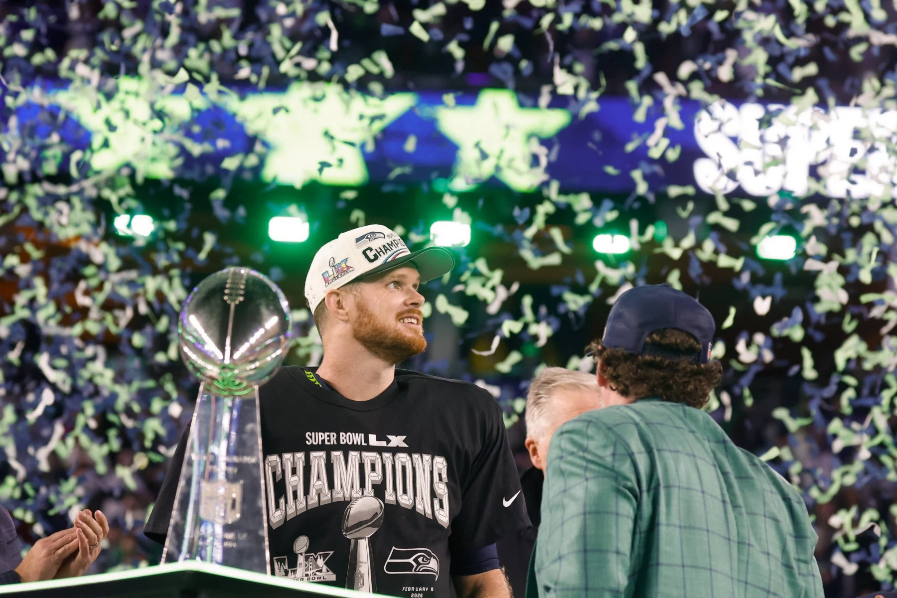 Sam Darnold - Seattle Seahawks - Ein Football-Spieler, der ein "Champion"-Shirt und eine Kappe trägt, steht neben der Super-Bowl-Trophäe, während Konfetti herabfällt. Um ihn herum auf dem Spielfeld feiern andere den Sieg der Seattle Seahawks. Alt-Text wurde KI-generiert.