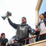 Sam Darnold hält eine Trophäe auf einem Umzugswagen in Seattle hoch, umgeben von anderen Personen, von denen einige Fotos machen und Getränke in der Hand halten, um den Super-Bowl-Sieg der Seahawks zu feiern. Alt-Text wurde KI-generiert.