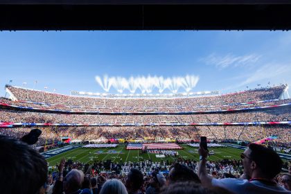 Ein volles Football-Stadion mit einer großen Menschenmenge, Feuerwerk am Himmel und die US-Flagge auf dem Spielfeld bilden die Kulisse für den Super Bowl 60, wo die Vorfreude auf einen möglichen Auftritt von Bad Bunny und einen weiteren Rekordmarken-Moment wächst. Alt-Text wurde KI-generiert.