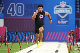 Kenyon Sadiq - Ein Athlet in einem schwarzen Outfit mit der Aufschrift "23 TE" absolviert beim NFL Scouting Combine 2026 einen 40-Yard-Lauf, umgeben von Zeitmessgeräten auf einem Hallenfeld neben anderen herausragenden Tight Ends und Defensive Backs. Alt-Text wurde KI-generiert.