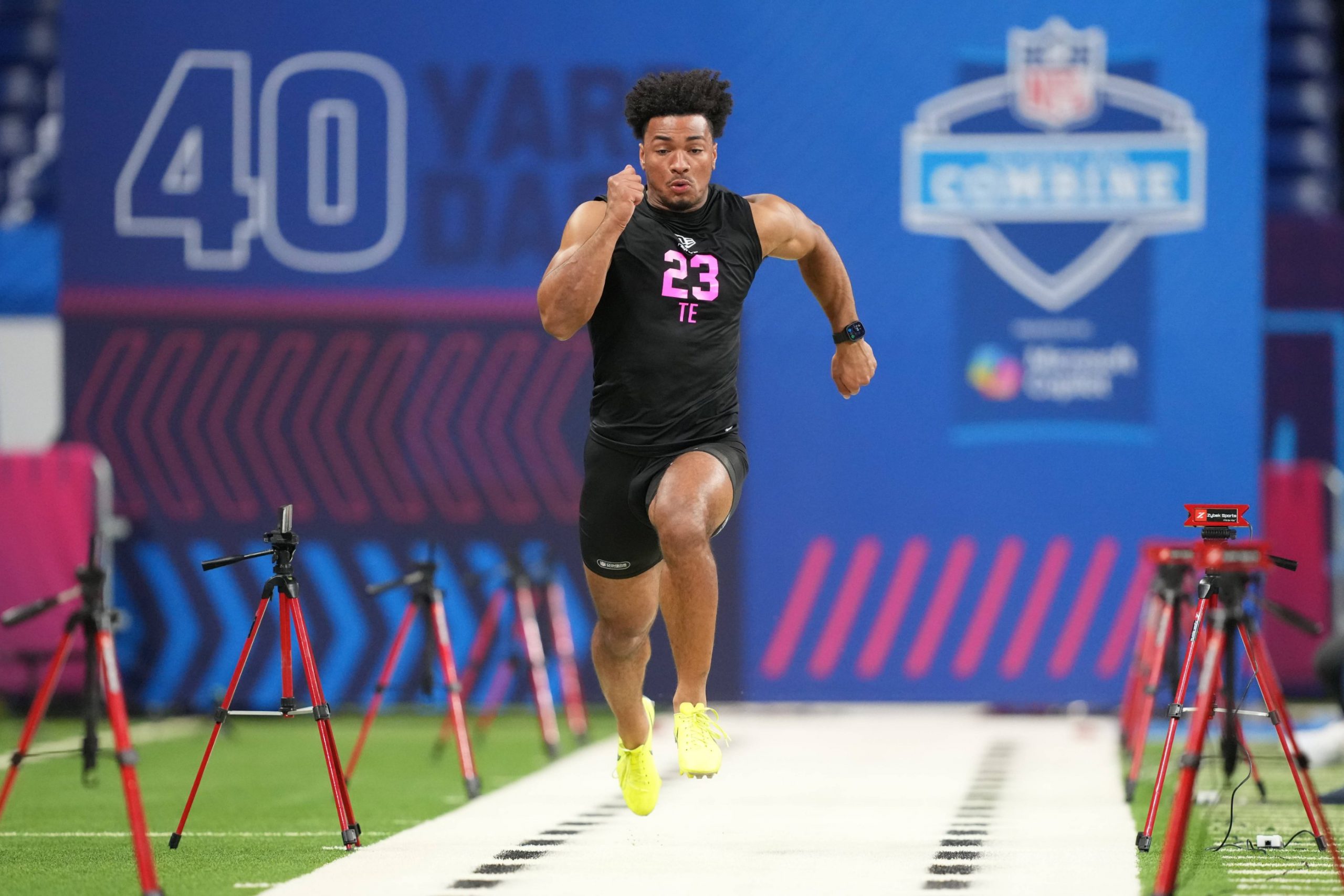 Kenyon Sadiq - Ein Athlet in einem schwarzen Outfit mit der Aufschrift "23 TE" absolviert beim NFL Scouting Combine 2026 einen 40-Yard-Lauf, umgeben von Zeitmessgeräten auf einem Hallenfeld neben anderen herausragenden Tight Ends und Defensive Backs. Alt-Text wurde KI-generiert.