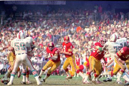 Ein laufendes Footballspiel zwischen dem Washingtoner Team in roten Trikots und den Miami Dolphins in weißen Trikots, das an den legendären Hall of Fame-QB Sonny Jurgensen erinnert, während die Spieler aktiv auf dem Spielfeld sind. Alt-Text wurde KI-generiert.