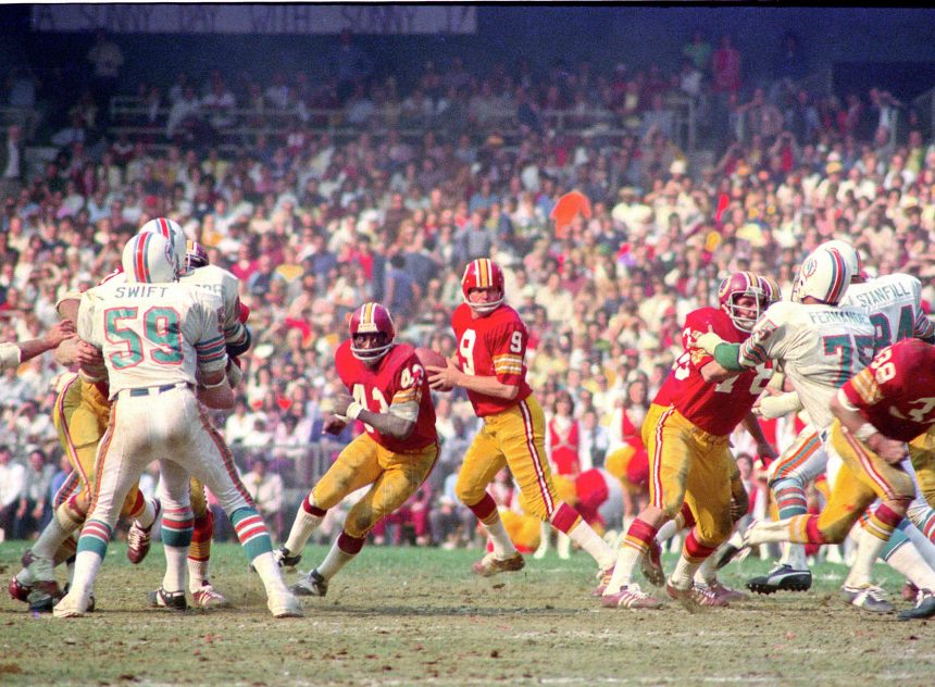 Ein laufendes Footballspiel zwischen dem Washingtoner Team in roten Trikots und den Miami Dolphins in weißen Trikots, das an den legendären Hall of Fame-QB Sonny Jurgensen erinnert, während die Spieler aktiv auf dem Spielfeld sind. Alt-Text wurde KI-generiert.