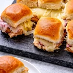 Eine Schiefertafel mit Hawaiian Pizza Sliders - gebackener Schinken und Käse auf weichen Brötchen, umstritten wegen ihres süß-salzigen Geschmacks; ein paar verlockende Sliders liegen auf einer weißen Platte im Vordergrund. Alt-Text wurde KI-generiert.