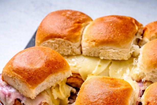Eine Schiefertafel mit Hawaiian Pizza Sliders - gebackener Schinken und Käse auf weichen Brötchen, umstritten wegen ihres süß-salzigen Geschmacks; ein paar verlockende Sliders liegen auf einer weißen Platte im Vordergrund. Alt-Text wurde KI-generiert.