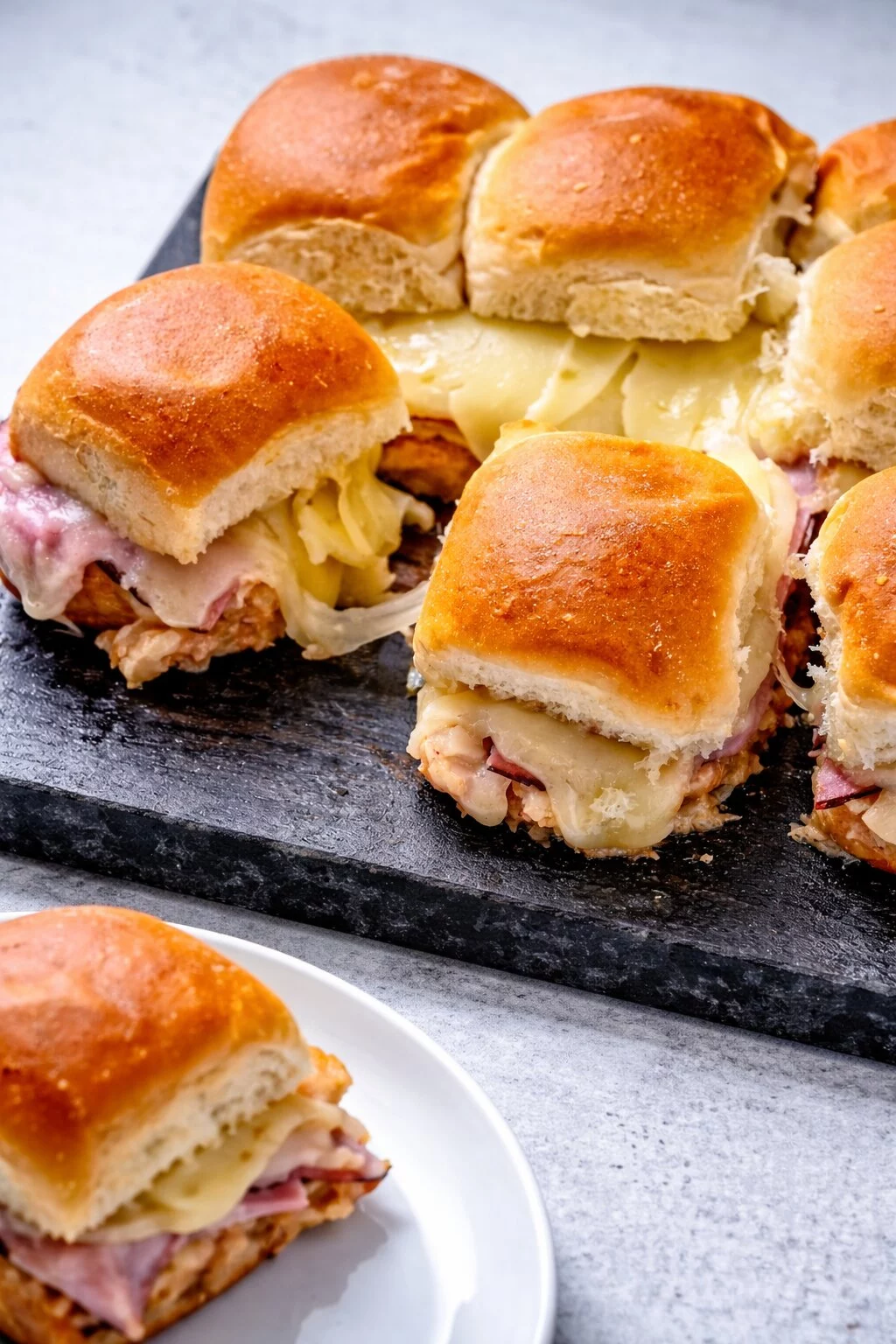 Eine Schiefertafel mit Hawaiian Pizza Sliders - gebackener Schinken und Käse auf weichen Brötchen, umstritten wegen ihres süß-salzigen Geschmacks; ein paar verlockende Sliders liegen auf einer weißen Platte im Vordergrund. Alt-Text wurde KI-generiert.