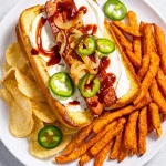 Ein Teller mit einem Seattle-Style Hot Dog mit Sauerrahm, gegrillten Zwiebeln, Jalapeño-Scheiben und Barbecue-Soße, serviert mit Süßkartoffelpommes und Kartoffelchips - perfekt für den Super Bowl-Snack. Alt-Text wurde KI-generiert.