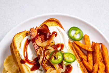 Ein Teller mit einem Seattle-Style Hot Dog mit Sauerrahm, gegrillten Zwiebeln, Jalapeño-Scheiben und Barbecue-Soße, serviert mit Süßkartoffelpommes und Kartoffelchips - perfekt für den Super Bowl-Snack. Alt-Text wurde KI-generiert.