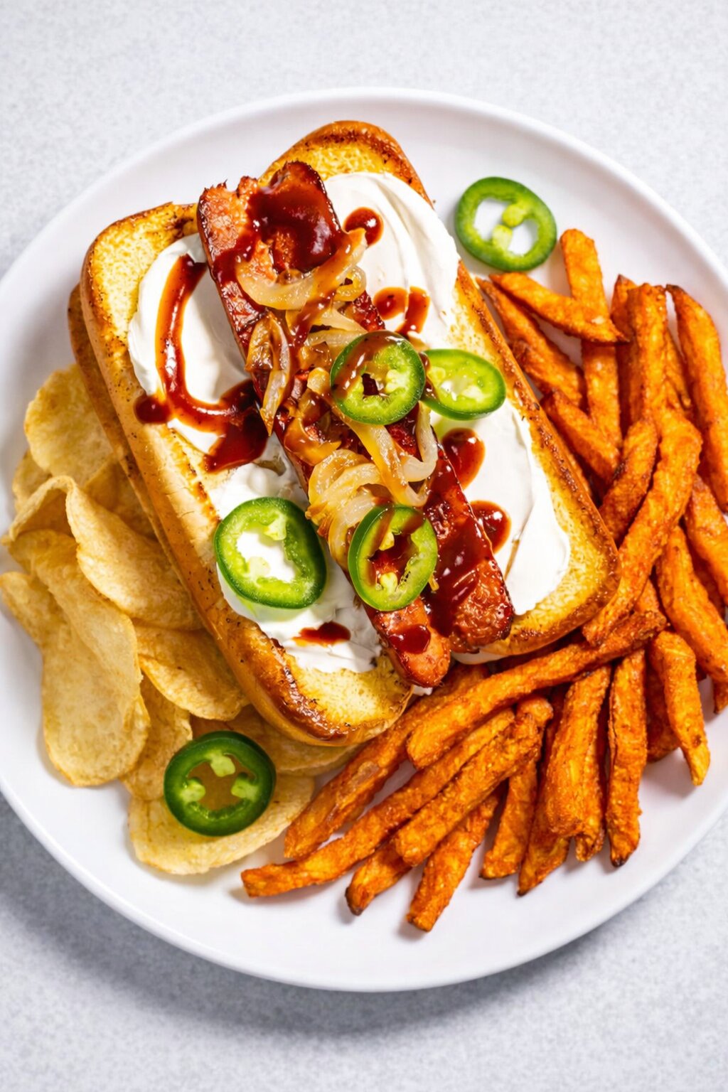 Ein Teller mit einem Seattle-Style Hot Dog mit Sauerrahm, gegrillten Zwiebeln, Jalapeño-Scheiben und Barbecue-Soße, serviert mit Süßkartoffelpommes und Kartoffelchips - perfekt für den Super Bowl-Snack. Alt-Text wurde KI-generiert.