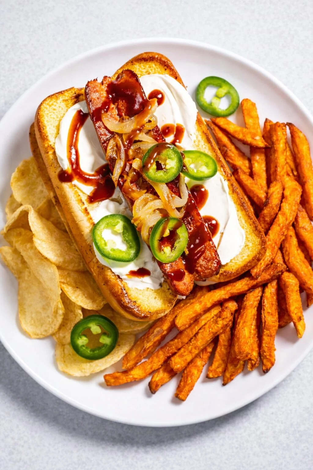 Ein Teller mit einem Seattle-Style Hot Dog mit Sauerrahm, gegrillten Zwiebeln, Jalapeño-Scheiben und Barbecue-Soße, serviert mit Süßkartoffelpommes und Kartoffelchips - perfekt für den Super Bowl-Snack. Alt-Text wurde KI-generiert.