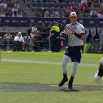 Flag Football Spieler in weiß-blauem Trikot mit Ball auf Spielfeld vor Zuschauern und Sicherheitskräften