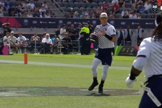 Flag Football Spieler in weiß-blauem Trikot mit Ball auf Spielfeld vor Zuschauern und Sicherheitskräften