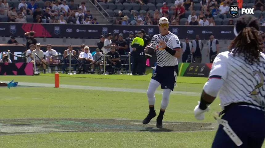 Flag Football Spieler in weiß-blauem Trikot mit Ball auf Spielfeld vor Zuschauern und Sicherheitskräften