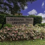 Stein mit Aufschrift Louisiana Tech University umgeben von bunten Pflanzen und grünen Büschen bei blauem Himmel