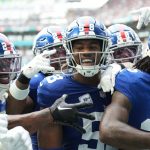 Eine Gruppe von New York Giants-Footballspielern, darunter Bobby Okereke in ihren charakteristischen blauen Trikots, feiert während eines Spiels gemeinsam auf dem Spielfeld. Alt-Text wurde KI-generiert.