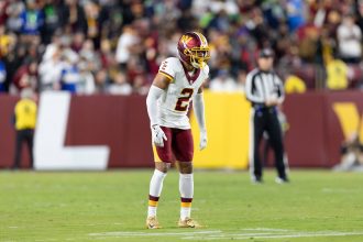 Marshon Lattimore - Ein Football-Spieler in einer weiß-bordeauxfarbenen Uniform der Commanders steht auf dem Spielfeld, bereit für das nächste Spiel, mit Zuschauern und Offiziellen im Hintergrund. Alt-Text wurde KI-generiert.