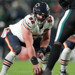 Drew Dalman - Chicago Bears-Spieler in weißer Uniform, Nummer 62, geht in die Hocke und bereitet sich darauf vor, den Ball während eines Spiels gegen eine Mannschaft in dunklen Uniformen zu werfen. Alt-Text wurde KI-generiert.