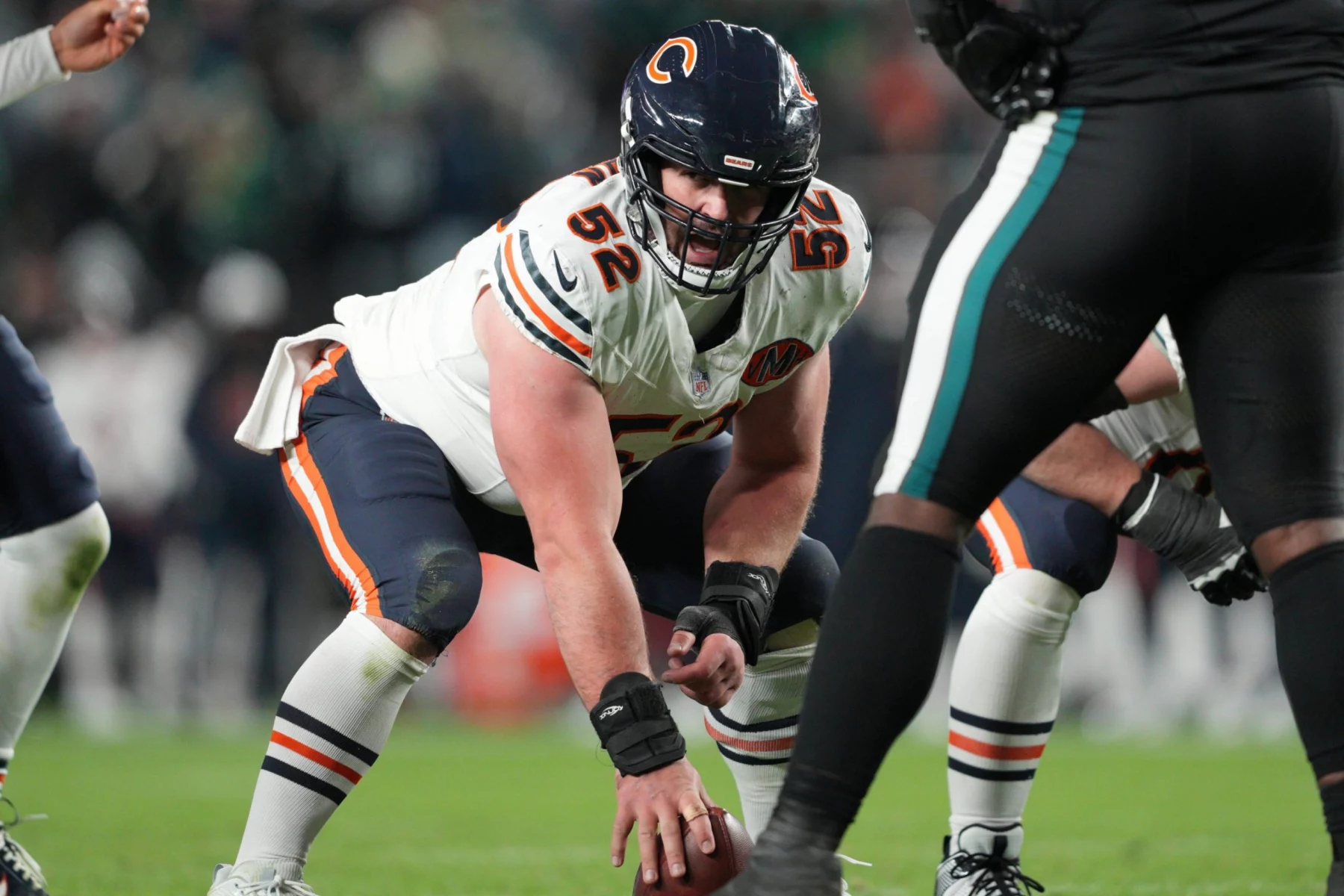 Drew Dalman - Chicago Bears-Spieler in weißer Uniform, Nummer 62, geht in die Hocke und bereitet sich darauf vor, den Ball während eines Spiels gegen eine Mannschaft in dunklen Uniformen zu werfen. Alt-Text wurde KI-generiert.