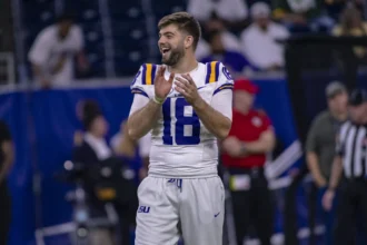 LSU-Quarterback Garrett Nussmeier, der die Nummer 16 trägt, steht auf dem Spielfeld und lächelt und klatscht, während im Hintergrund Zuschauer und Mitarbeiter zu sehen sind - ein Hoffnungsträger für den NFL-Draft 2026. Alt-Text wurde KI-generiert.