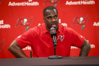 Lavonte David in rotem Tampa Bay Buccaneers Poloshirt spricht bei Pressekonferenz vor rotem AdventHealth-Hintergrund
