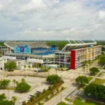 Luftaufnahme des Camping World Stadium in Orlando mit seinen mehrfarbigen Außenwänden, umgeben von Bäumen, Parkplätzen und nahe gelegenen Straßen unter einem teilweise bewölkten Himmel - Heimat spannender Veranstaltungen wie Jaguars-Spiele. Alt-Text wurde KI-generiert.