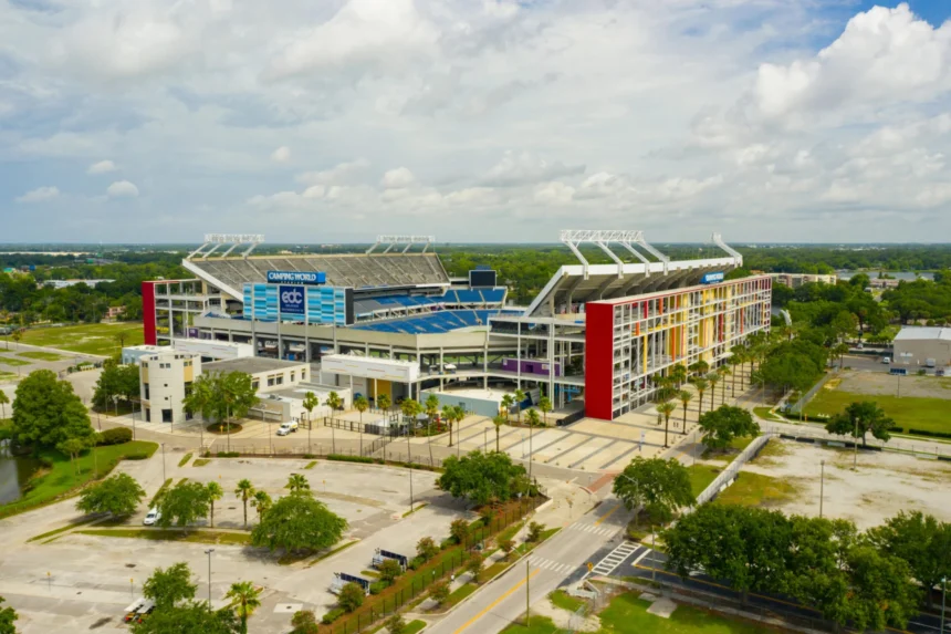 Luftaufnahme des Camping World Stadium in Orlando mit seinen mehrfarbigen Außenwänden, umgeben von Bäumen, Parkplätzen und nahe gelegenen Straßen unter einem teilweise bewölkten Himmel - Heimat spannender Veranstaltungen wie Jaguars-Spiele. Alt-Text wurde KI-generiert.