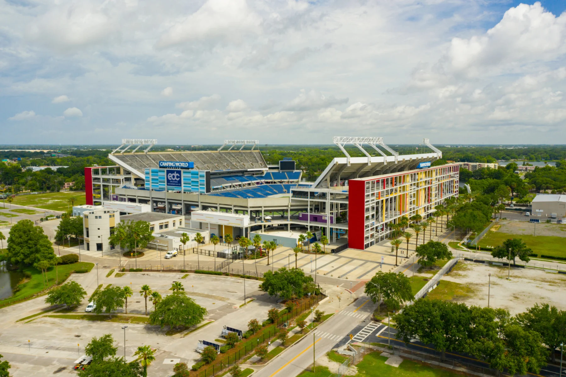 Luftaufnahme des Camping World Stadium in Orlando mit seinen mehrfarbigen Außenwänden, umgeben von Bäumen, Parkplätzen und nahe gelegenen Straßen unter einem teilweise bewölkten Himmel - Heimat spannender Veranstaltungen wie Jaguars-Spiele. Alt-Text wurde KI-generiert.