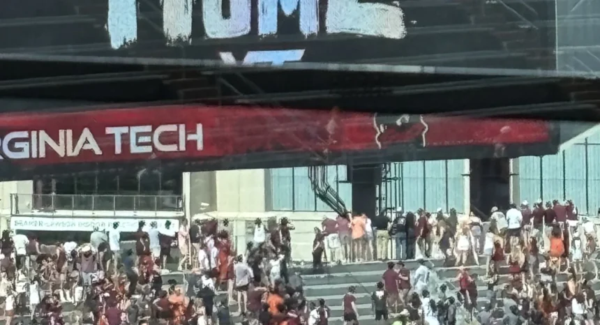 Vor einem Gebäude in der Nähe des Lane Stadium versammelt sich eine Menschenmenge, viele in kastanienbrauner Kleidung, unter einem "VIRGINIA TECH"-Banner und einem teilweise sichtbaren "HOME"-Schild darüber. Alt-Text wurde KI-generiert.