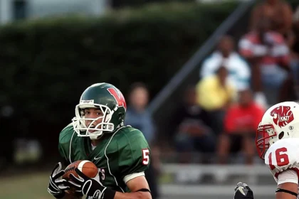 Ein Football-Spieler in einem grünen Trikot rennt mit dem Ball, während ein Spieler in einem rot-weißen Trikot aus Österreich ihn auf dem Spielfeld verfolgt und so den Geist des American Football und den wachsenden NFL-Hype einfängt. Alt-Text wurde KI-generiert.