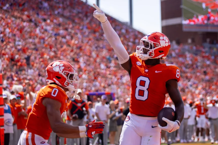 Zwei Clemson-Football-Spieler, darunter Adam Randall, feiern in orangefarbenen Trikots vor einer jubelnden Menge einen Touchdown; einer hält den Ball und zeigt nach oben - und unterstreicht damit sein Spielerprofil im Vorfeld des NFL Draft 2026. Alt-Text wurde KI-generiert.