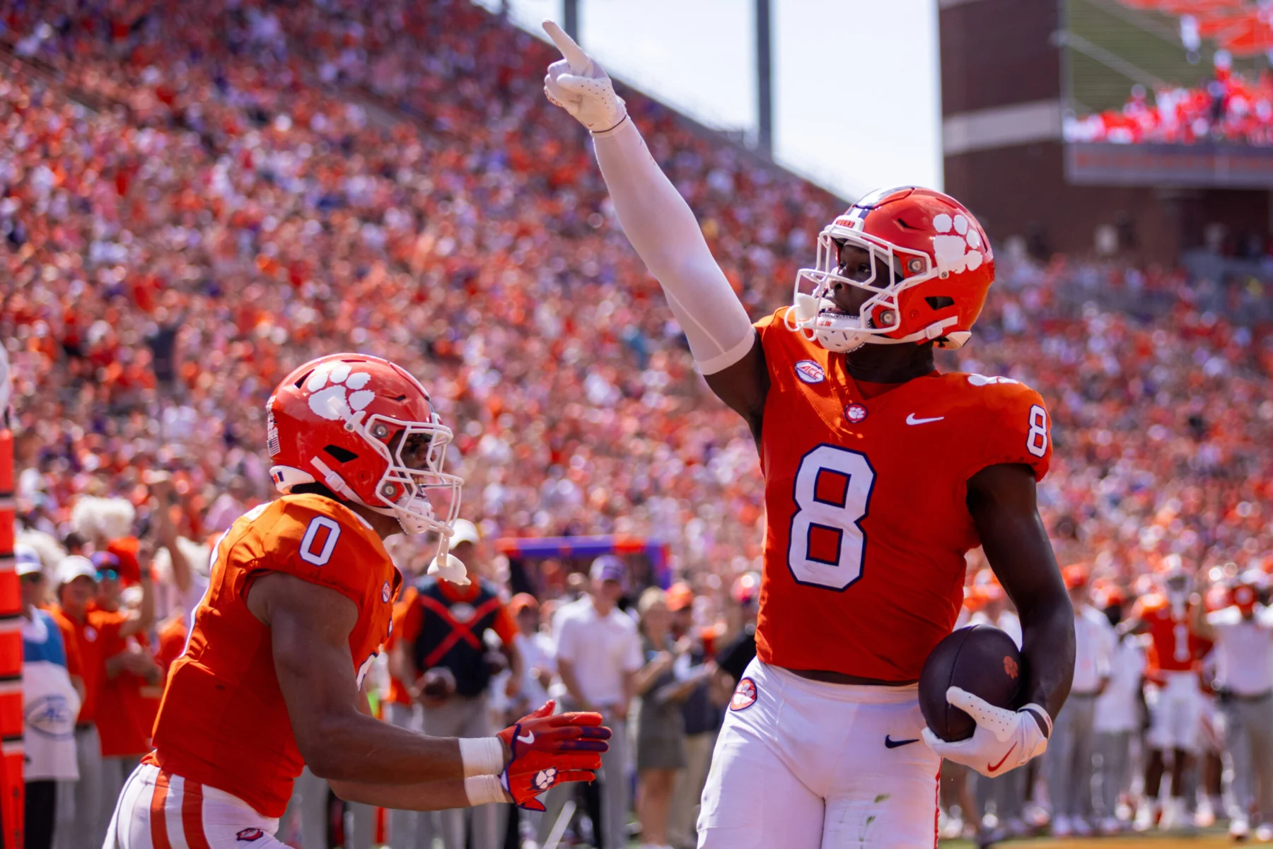 Zwei Clemson-Football-Spieler, darunter Adam Randall, feiern in orangefarbenen Trikots vor einer jubelnden Menge einen Touchdown; einer hält den Ball und zeigt nach oben - und unterstreicht damit sein Spielerprofil im Vorfeld des NFL Draft 2026. Alt-Text wurde KI-generiert.