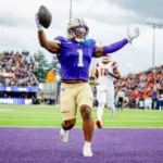 Ein College-Football-Spieler, Jonah Coleman, in einem lila Trikot läuft mit einem Football in die Endzone und feiert einen Touchdown, während die Gegner hinterherlaufen - und zeigt dabei Fähigkeiten, die sein NFL Draft 2026 Spielerprofil verbessern könnten. Alt-Text wurde KI-generiert.