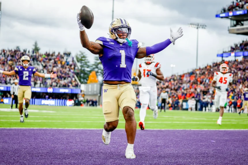 Ein College-Football-Spieler, Jonah Coleman, in einem lila Trikot läuft mit einem Football in die Endzone und feiert einen Touchdown, während die Gegner hinterherlaufen - und zeigt dabei Fähigkeiten, die sein NFL Draft 2026 Spielerprofil verbessern könnten. Alt-Text wurde KI-generiert.